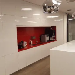 Biała, nowoczesna kuchnia z połyskiem, czerwony panel nad blatem, chromowane lampy wiszące i ekspres do kawy.