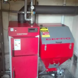 Czerwono-szary piec CO marki DEFRO z podajnikiem na ekogroszek, rury instalacji grzewczej, widoczne przyłącza i manometr, w tle ściana z widocznymi śladami użytkowania.