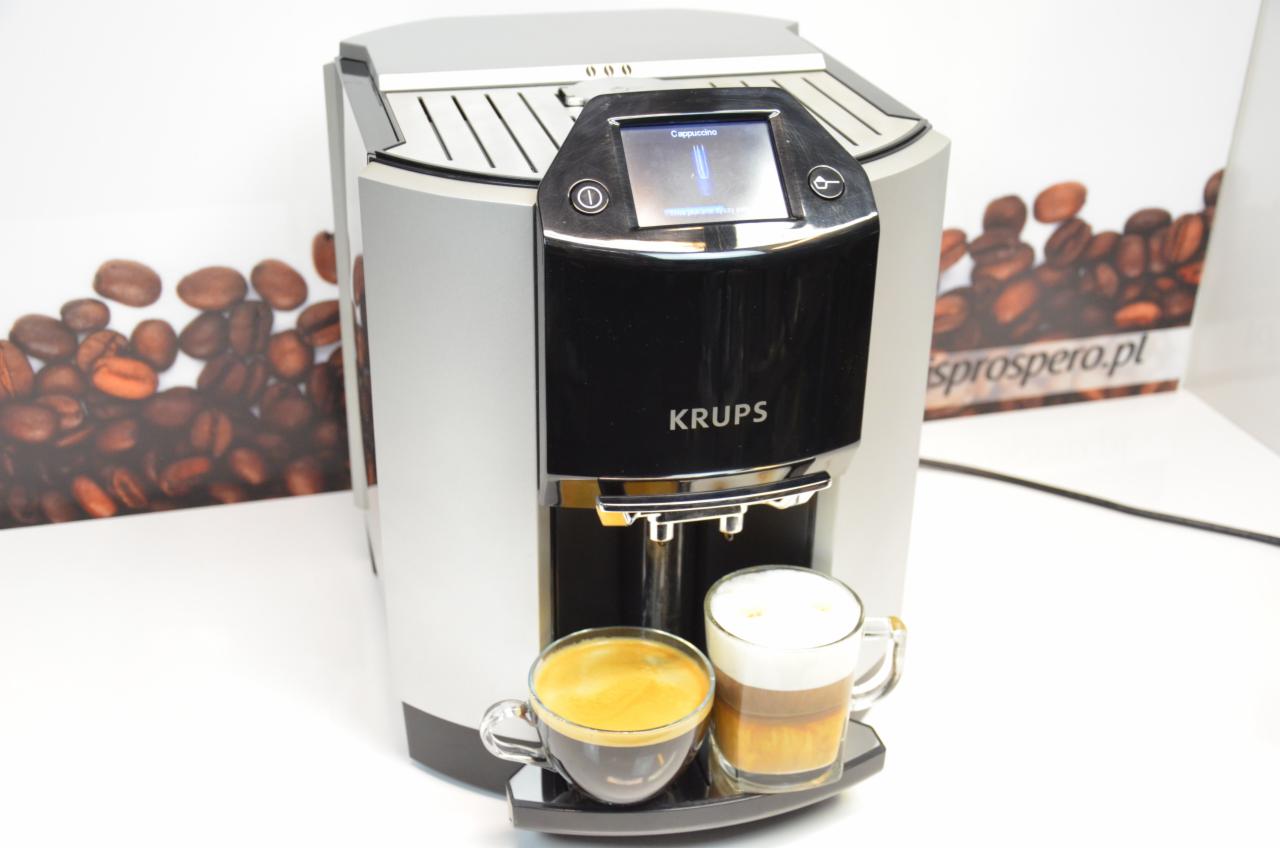 Srebrny ekspres do kawy Krups z dwoma szklanymi filiżankami: espresso i latte macchiato na czarnej podstawce, na tle reklamy z ziarnami kawy.