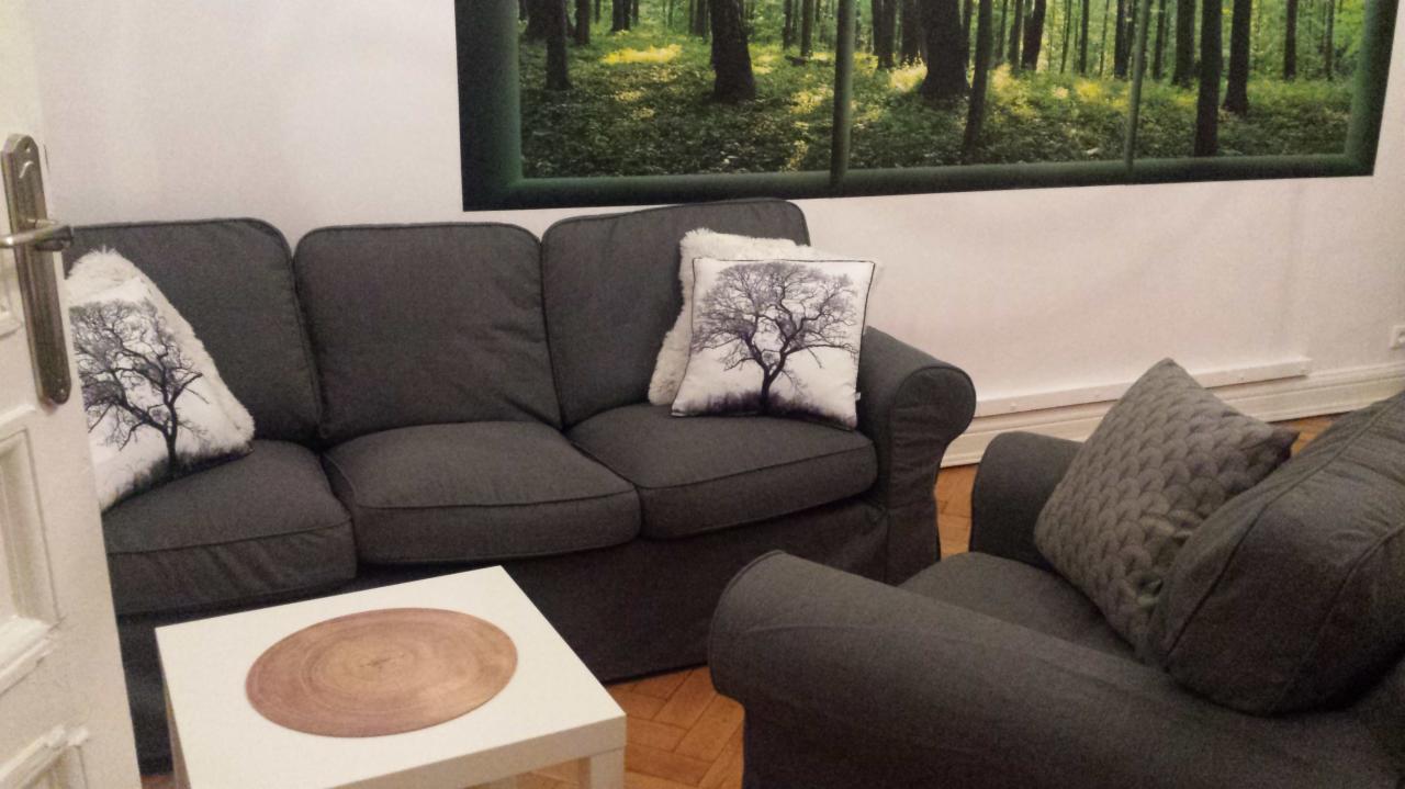 Szary sofa i fotel z poduszkami z motywem drzewa w pokoju z obrazem lasu na ścianie, widok zza uchylonych drzwi.