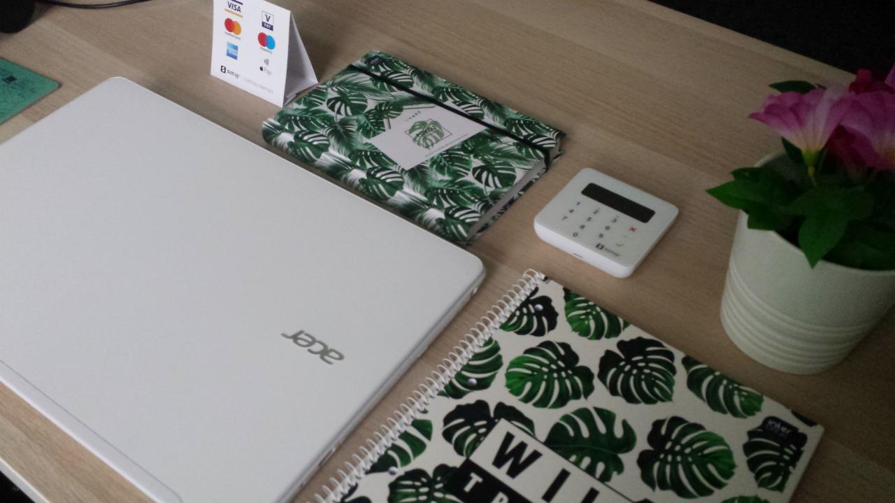 Jasny biurkowy układ z białym laptopem Acer, dwoma notatnikami z motywem liści monstery, terminalem płatniczym, ulotką akceptowanych kart i doniczką z różowymi kwiatami.