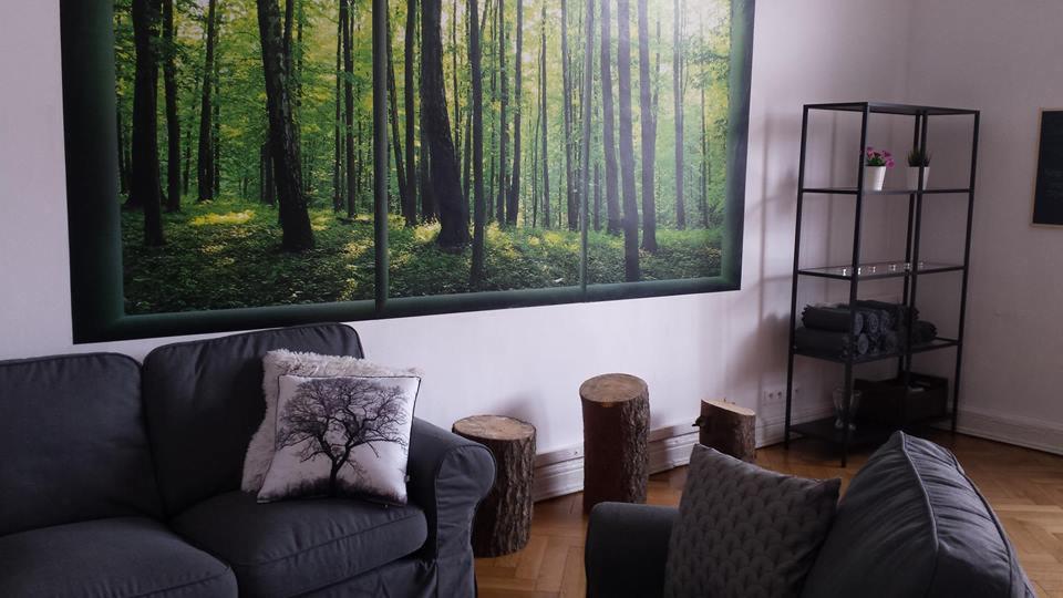 Wnętrze gabinetu z szarą sofą, poduszką z motywem drzewa, drewnianymi pufami i fototapetą z widokiem na las.