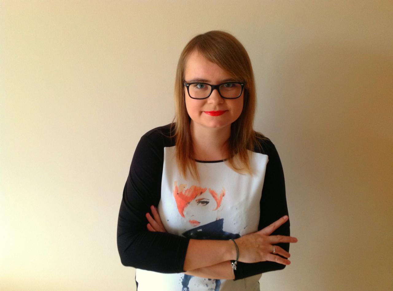 Portret kobiety w okularach z czerwonymi ustami, ubranej w koszulkę z nadrukiem postaci, stojącej na tle jasnej ściany.