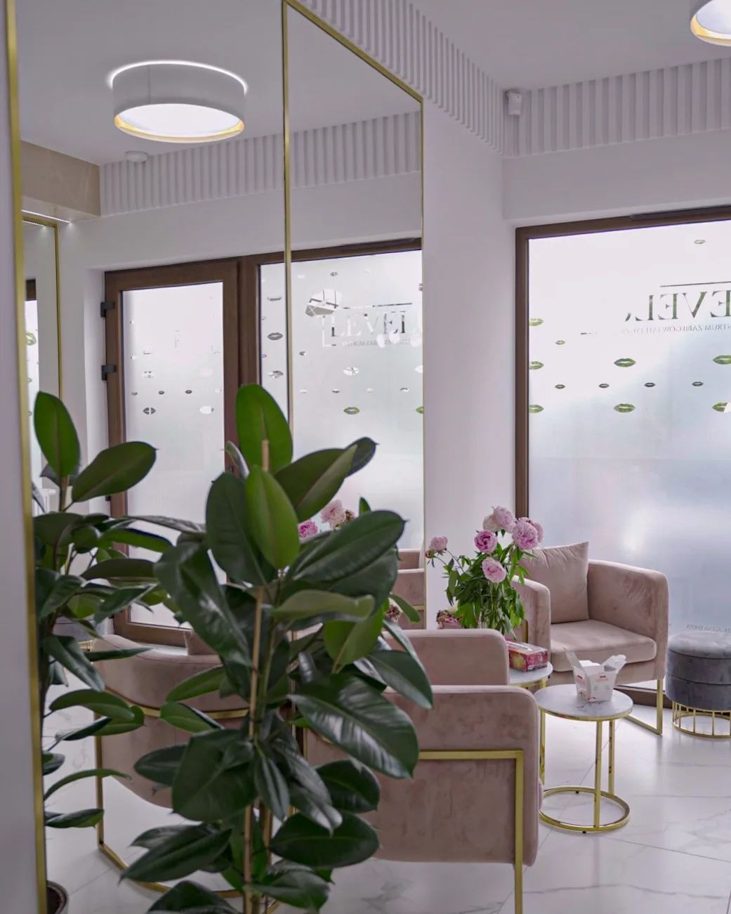 Eleganckie wnętrze poczekalni z marmurową podłogą, pluszowymi fotelami w odcieniach różu, stolikami kawowymi ze złotymi nogami i dużym lustrem w złotej ramie, dekorowane kwiatami i roślinami...