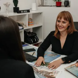 Uśmiechnięta kobieta w czarnej bluzce prezentuje katalog z ofertami wycieczek klientowi przy biurku z mapą świata.