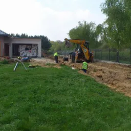 Prace ziemne koparką przy nowo budowanym domu jednorodzinnym, widoczni pracownicy w kamizelkach odblaskowych, wykopany rów wzdłuż ogrodzenia z siatki.
