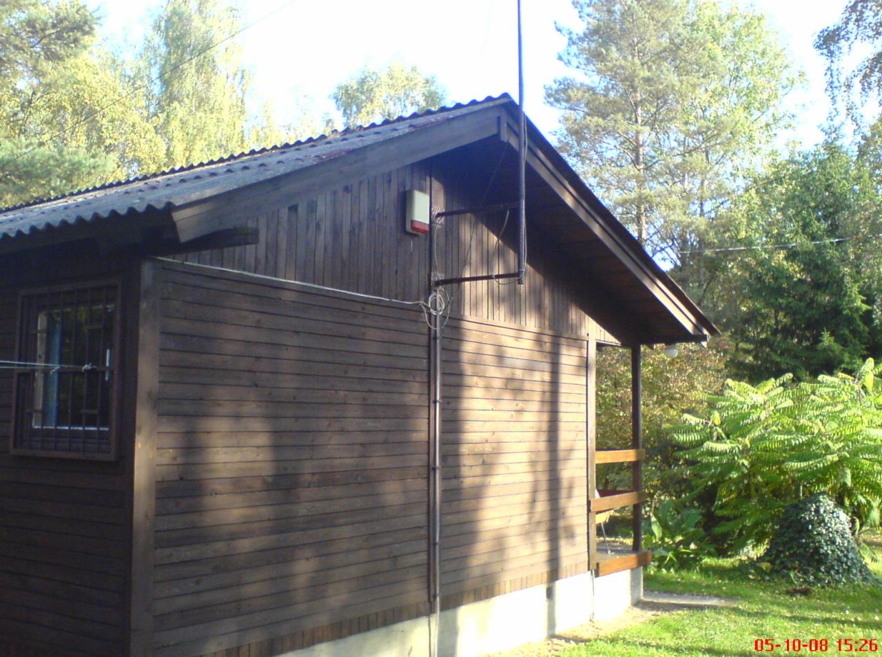 Brązowy drewniany budynek z zainstalowanym systemem odgromowym, widoczna pionowa rura i antena na dachu oraz alarm na ścianie, otoczony zielenią.