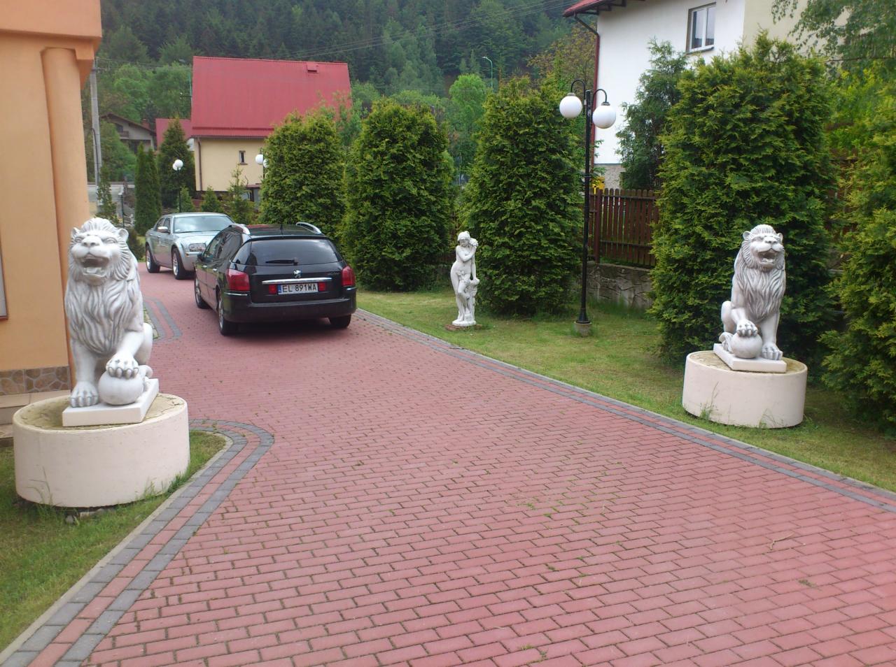 Posesja z brukowanym podjazdem, na którym stoją dwa samochody; po bokach podjazdu, na trawniku, ustawione są rzeźby lwów na postumentach i rzeźba kobiety.