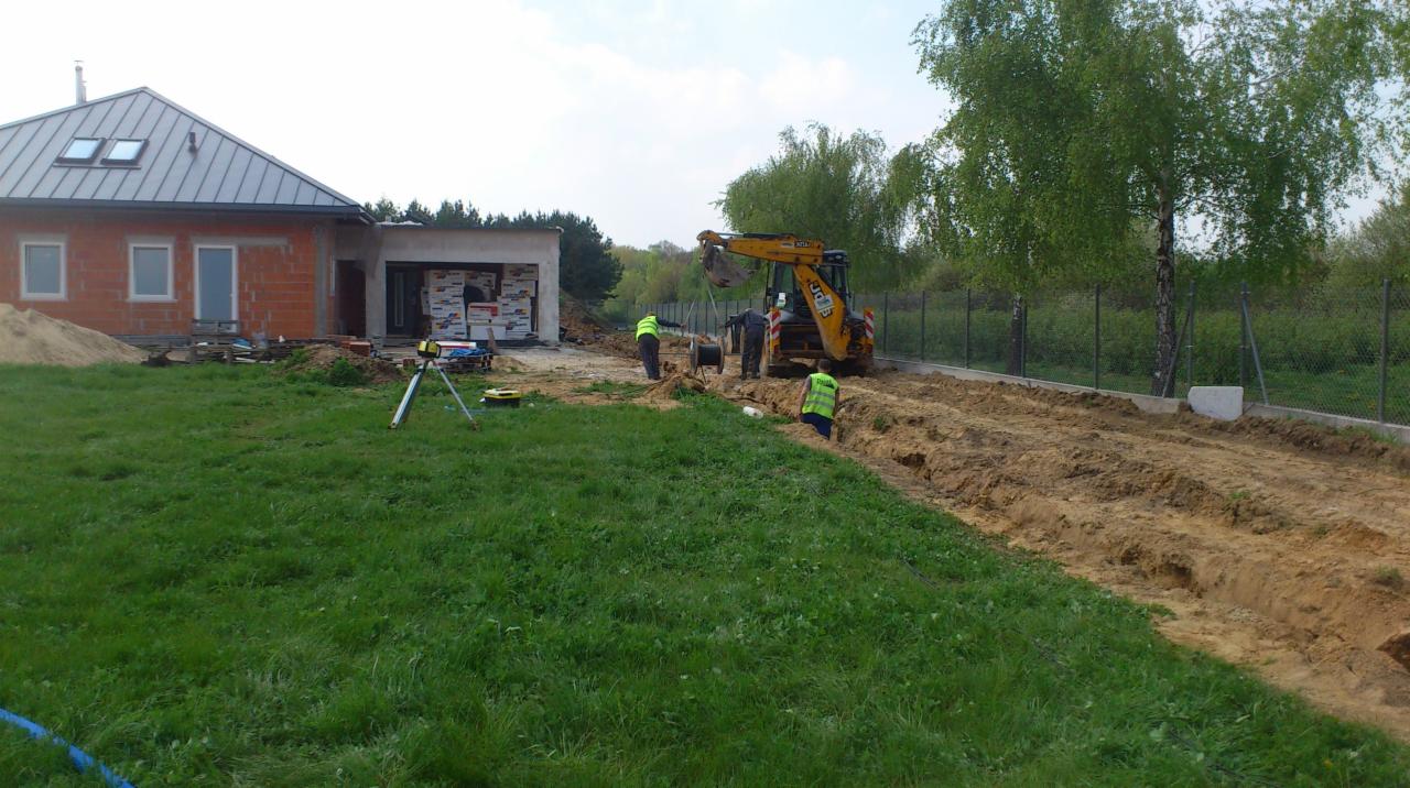 Prace ziemne koparką przy nowo budowanym domu jednorodzinnym, widoczni pracownicy w kamizelkach odblaskowych, wykopany rów wzdłuż ogrodzenia z siatki.