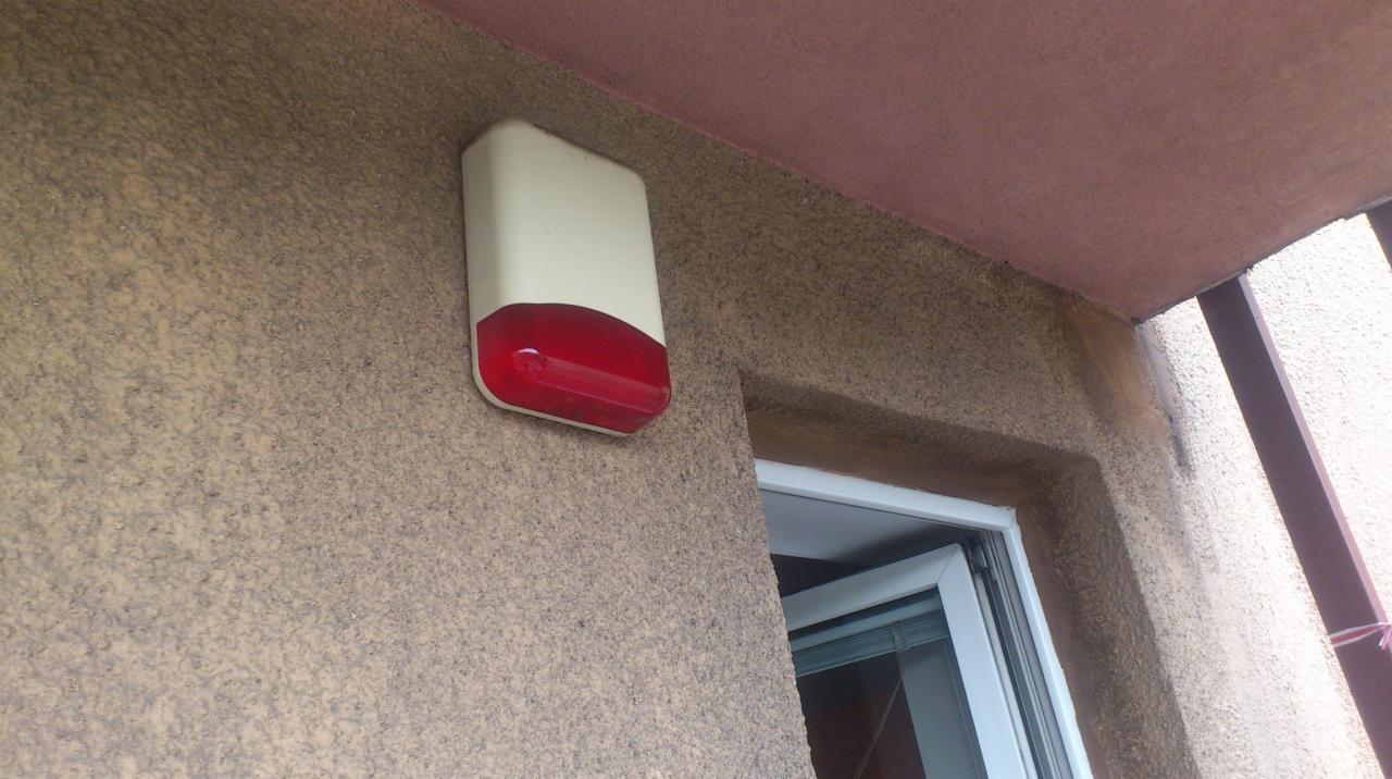 Zewnętrzna obudowa alarmowa z czerwoną lampą błyskową, zamontowana na chropowatej, brązowej elewacji budynku obok białej ramy okiennej.
