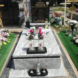 Granitowy pomnik z czarną tablicą, ozdobiony kwiatami, lampionami i krzyżem; widoczny portret zmarłego i inskrypcja na tablicy; otoczenie cmentarza z zielenią i innymi grobami.