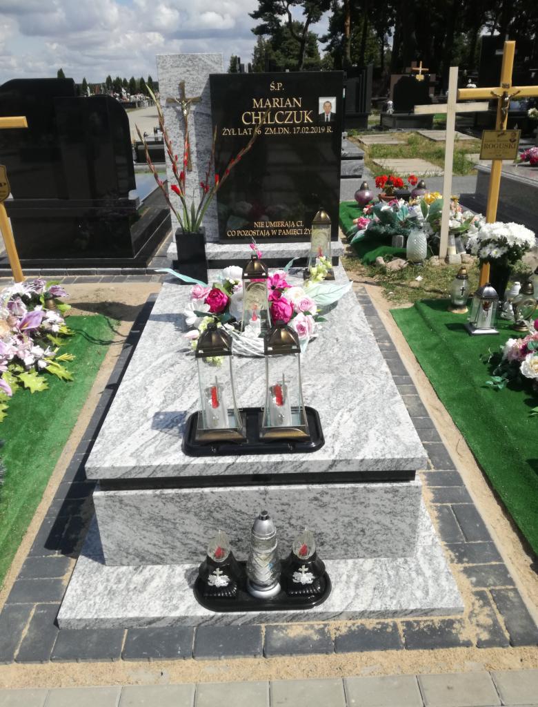 Granitowy pomnik z czarną tablicą, ozdobiony kwiatami, lampionami i krzyżem; widoczny portret zmarłego i inskrypcja na tablicy; otoczenie cmentarza z zielenią i innymi grobami.