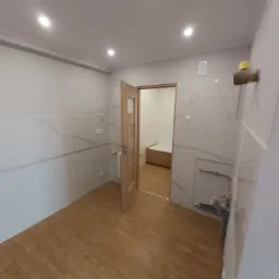 Wykończone wnętrze łazienki z drewnopodobną podłogą i płytkami imitującymi marmur na ścianach, widoczne instalacje hydrauliczne i otwarta drewniana rama drzwi prowadząca do kolejnej łazienki z wanną.