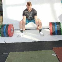 Mężczyzna w sportowym stroju wykonuje martwy ciąg ze sztangą obciążoną kolorowymi talerzami na tle białej ściany.