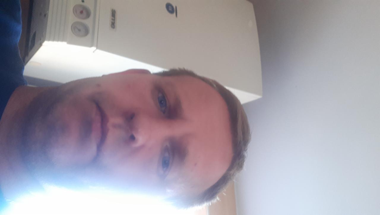 Mężczyzna robiący selfie na tle białego pieca gazowego marki ELLUS, w jasnym pomieszczeniu. Zdjęcie obrócone.