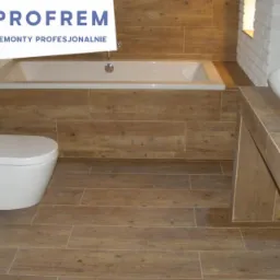 Nowoczesna łazienka z drewnopodobnymi płytkami na podłodze i ścianach, białą armaturą (umywalki, toaleta podwieszana, wanna) oraz fragment logo firmy w lewym górnym rogu.