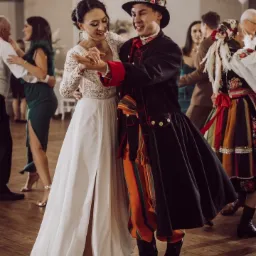 Para młoda w tańcu, on w stroju ludowym, ona w białej sukni z rozcięciem, na parkiecie w Radomsku, w tle goście i dekoracje.