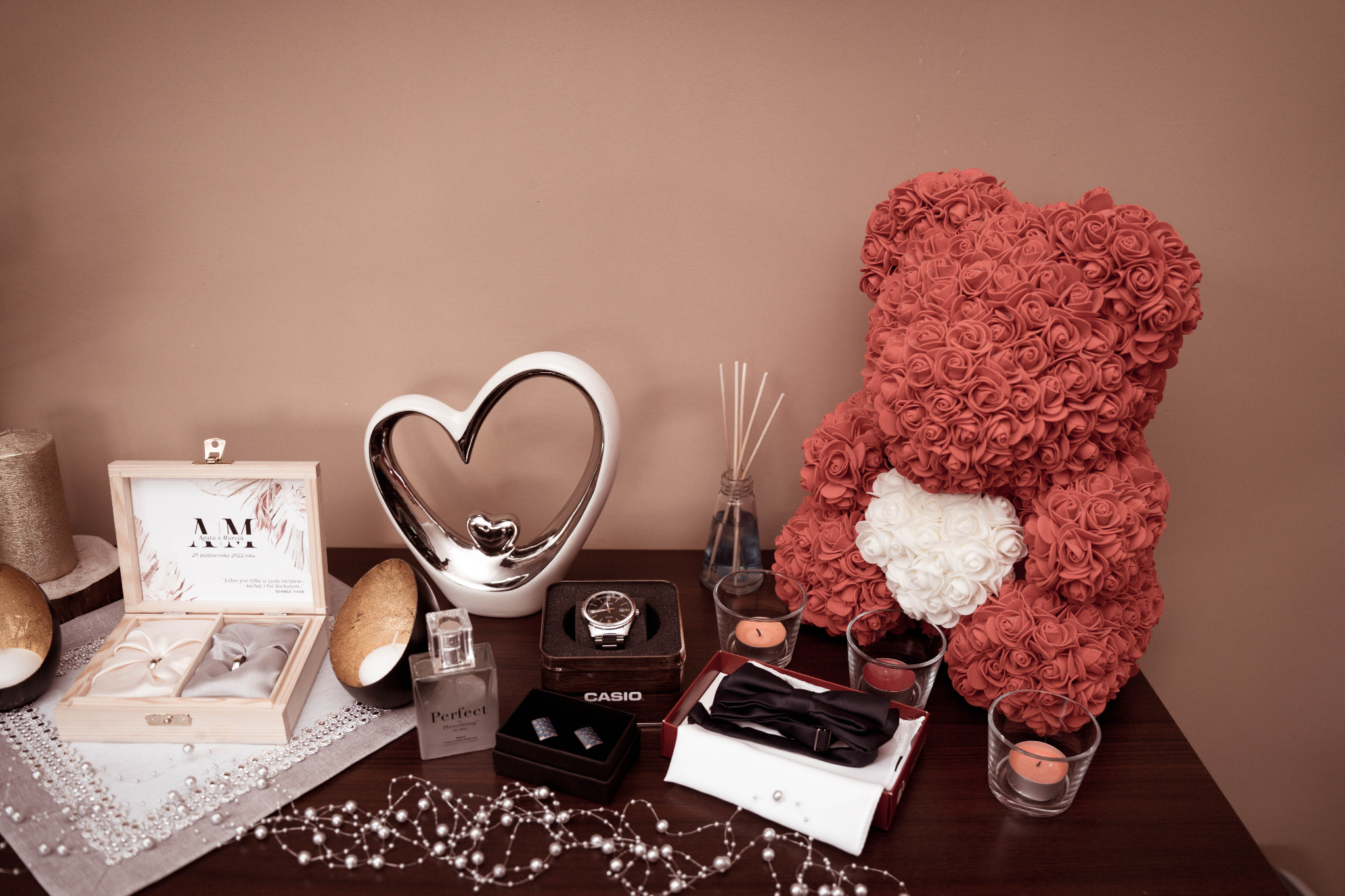 Kompozycja ślubna na stole: drewniane pudełko na obrączki z grawerem, pluszowy miś z róż, zegarek, muszka, spinki do mankietów, świece i dekoracyjne serce.
