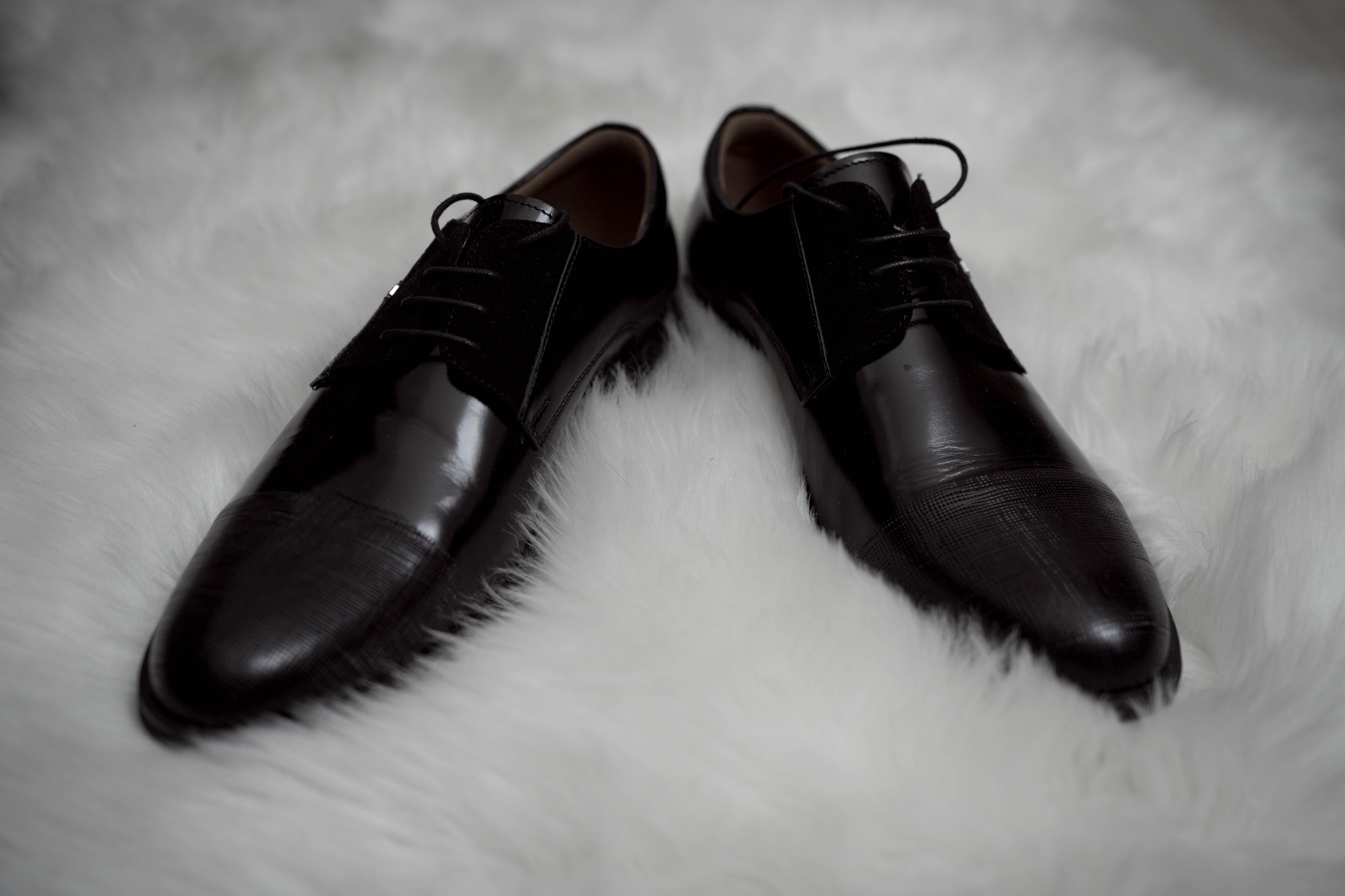 Para eleganckich, czarnych, lakierowanych męskich butów z delikatnym wzorem na czubkach, leżących na białym, puszystym dywanie.