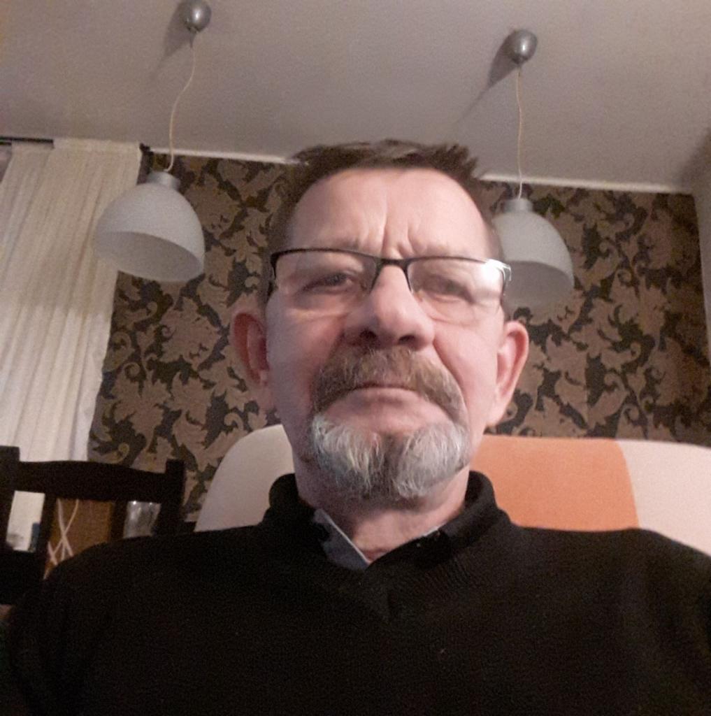 Portret mężczyzny z okularami i siwą brodą na tle pokoju z wzorzystą tapetą i lampami wiszącymi.