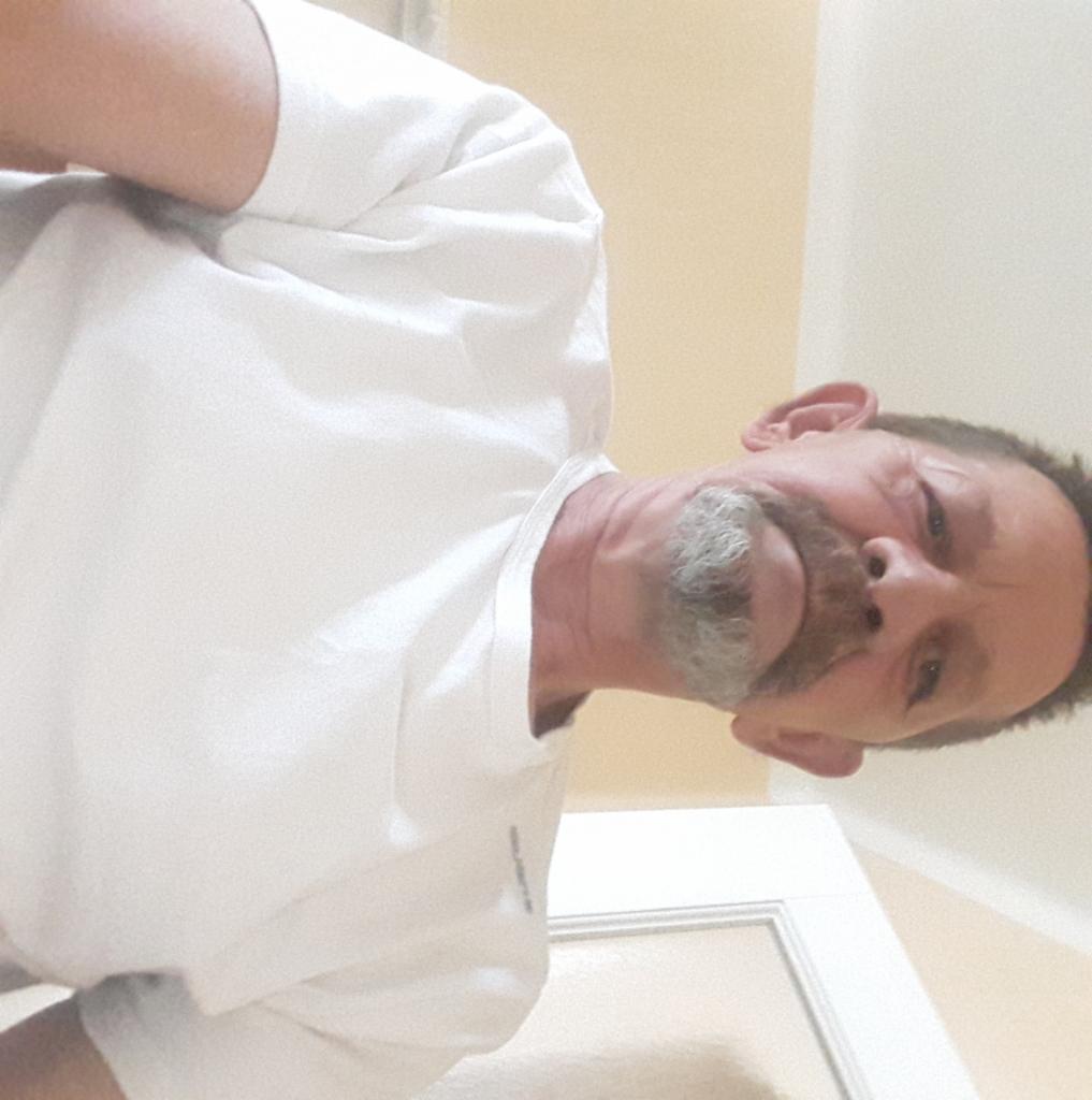 Mężczyzna w białej koszulce, z siwiejącą brodą, patrzy w dół, z perspektywy żabiej, na tle beżowej ściany i białych drzwi.