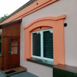 Odnowiona elewacja domu z oknem w ozdobnej, pomarańczowej ramie, drewnianym gankiem i kostką brukową przed wejściem.