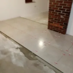 Fragment podłogi z ułożonymi płytkami w jasnym odcieniu, z użyciem czerwonych krzyżyków dystansowych, widoczna surowa wylewka podłogowa oraz fragment ściany z cegły klinkierowej w tle.