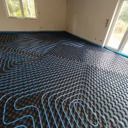 Niebieskie rurki ogrzewania podłogowego ułożone na czarnej macie w surowym pomieszczeniu z oknami i taśmą zabezpieczającą przy ścianach.