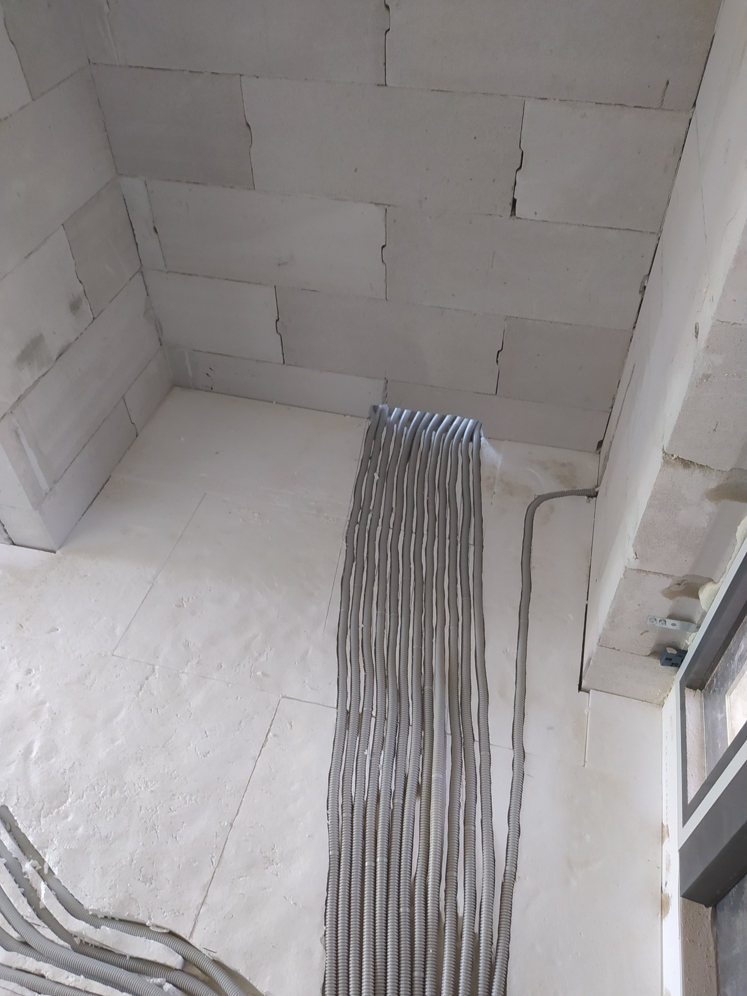 Szare, karbowane rury instalacyjne prowadzone pionowo w bruzdach w białej ścianie z betonu komórkowego, widoczne fragmenty okna i sufitu.