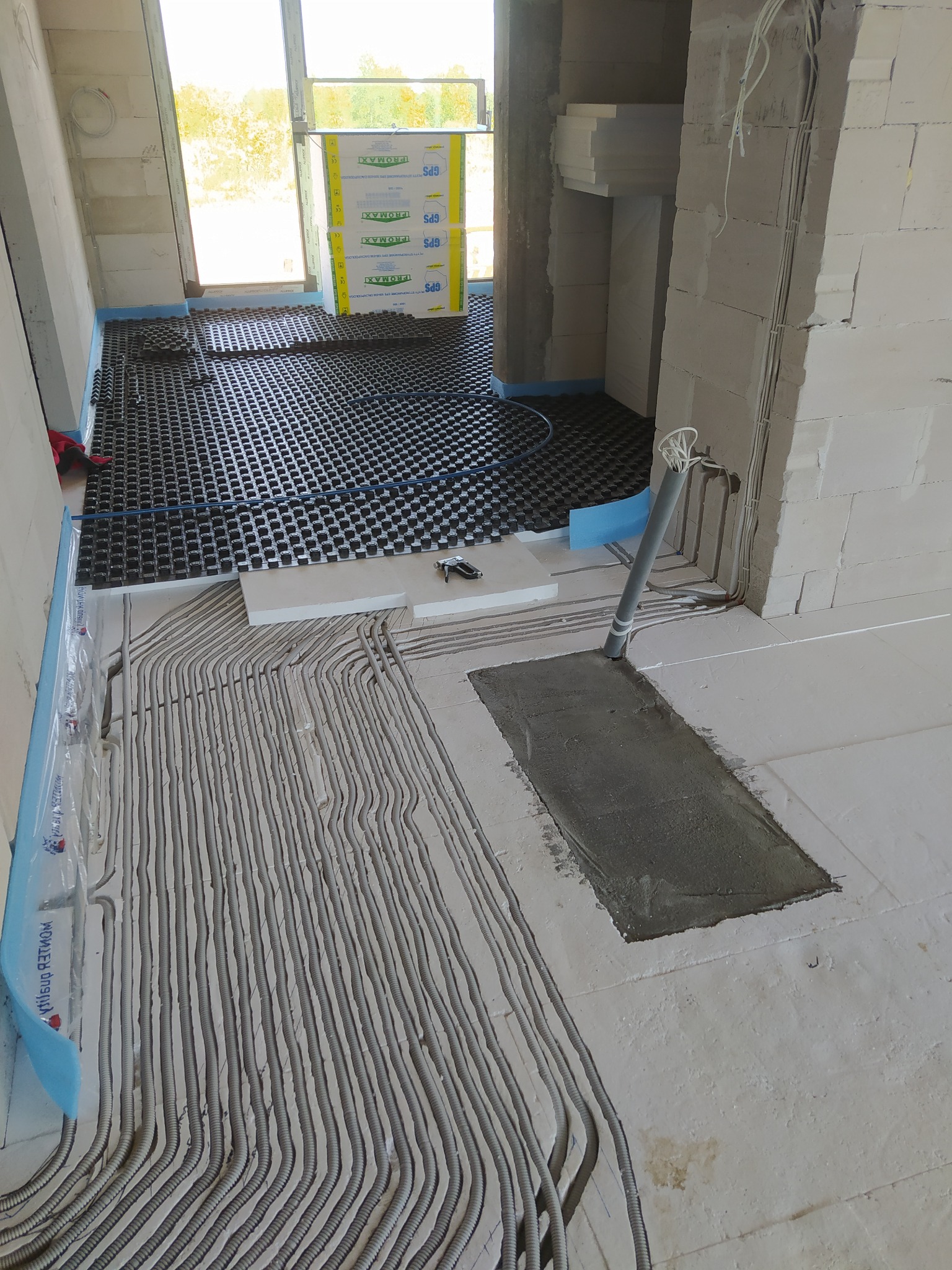 Instalacja ogrzewania podłogowego w surowym stanie, widoczne rury grzewcze ułożone na izolacji, przygotowane do zalania wylewką, z widocznym pionem rur. Częściowo ułożona mata systemowa.