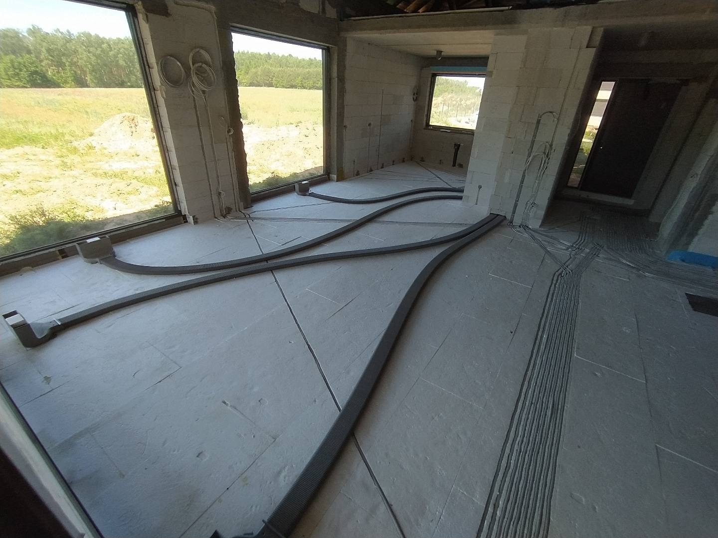 Wnętrze surowego domu w trakcie instalacji ogrzewania podłogowego, widoczne rury rozprowadzone na izolacji podłogowej, wystające przewody elektryczne ze ścian, duże okna z widokiem na pole.