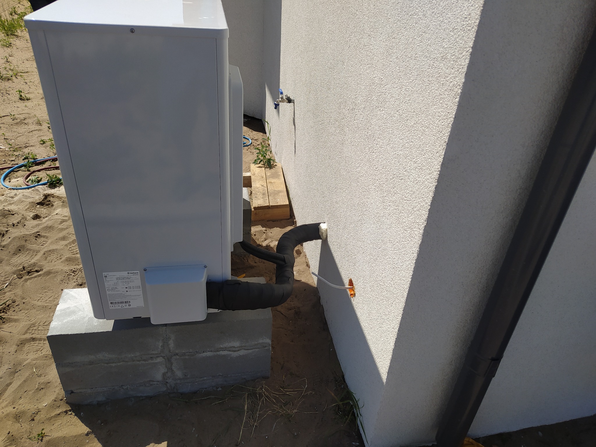 Zewnętrzna jednostka pompy ciepła marki Vaillant zamontowana na betonowym fundamencie przy ścianie budynku z widocznymi przewodami w izolacji termicznej.