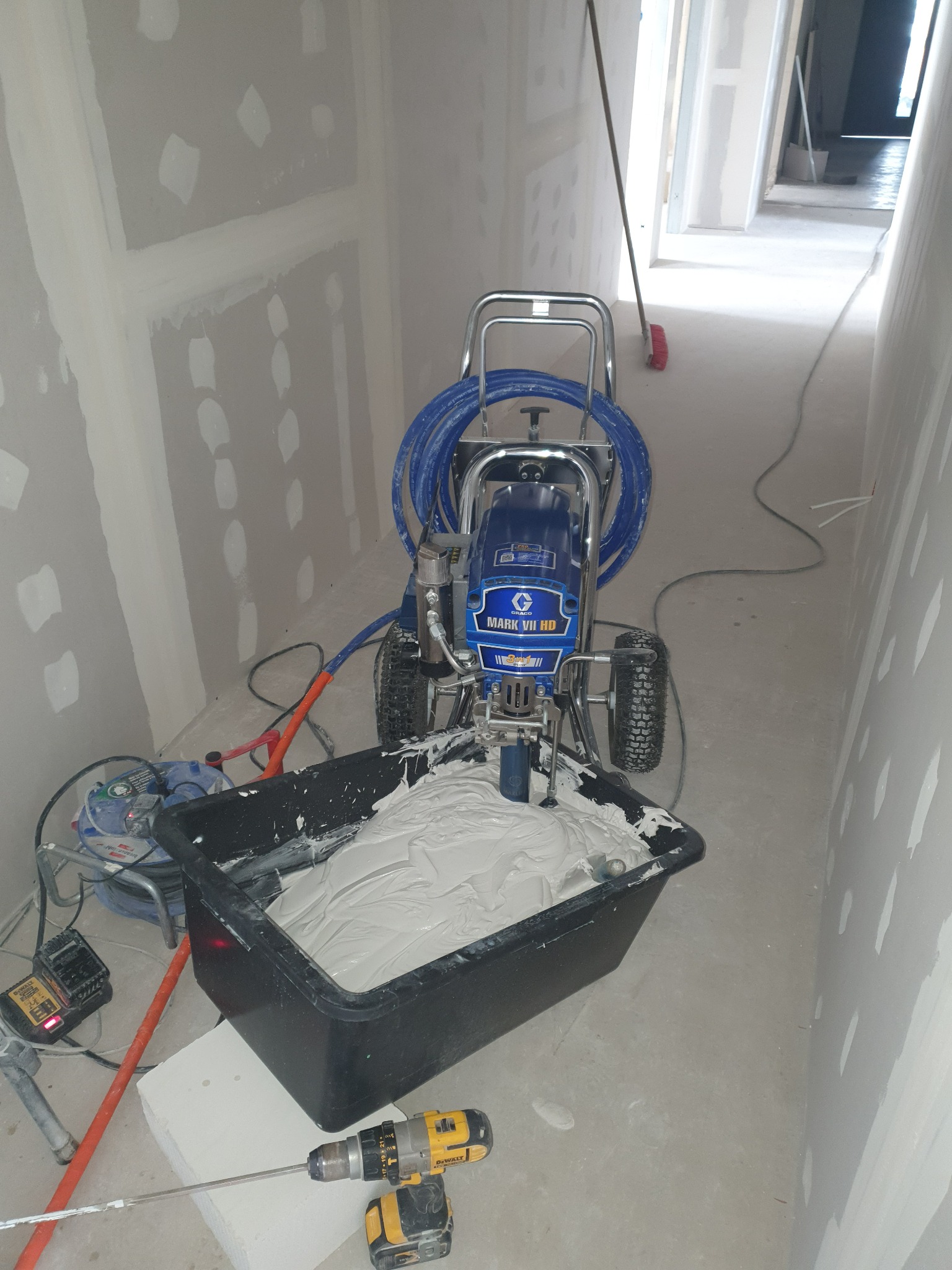 Agregat malarski Graco Mark VII HD z niebieskim wężem i czarnym pojemnikiem wypełnionym białą masą szpachlową, obok wiertarka DeWalt z mieszadłem, ściany przygotowane do gładzi