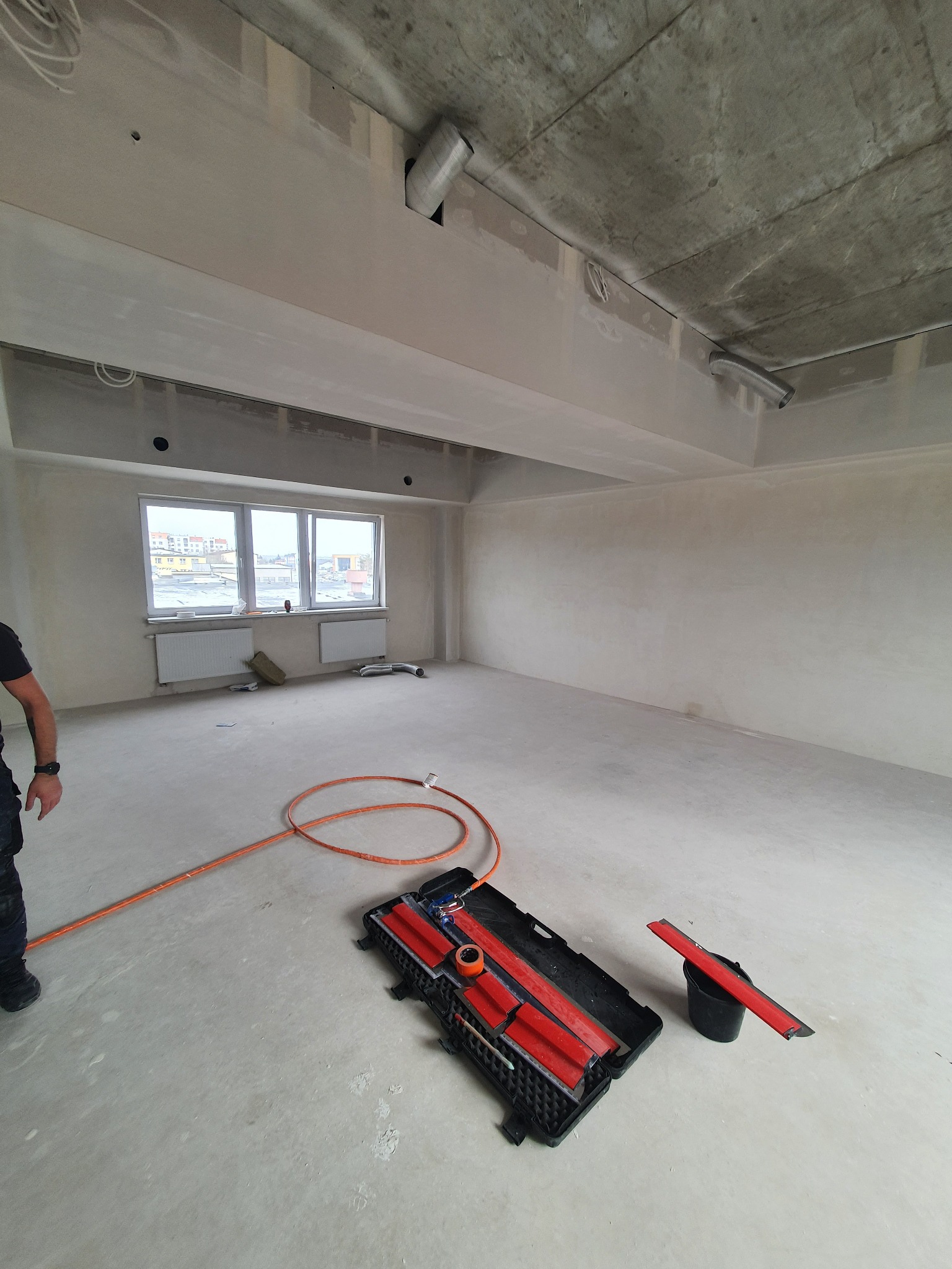 Wnętrze w stanie deweloperskim po gładziowaniu ścian, widoczne narzędzia do gładzi i szpachlowania, okno z widokiem na miasto.