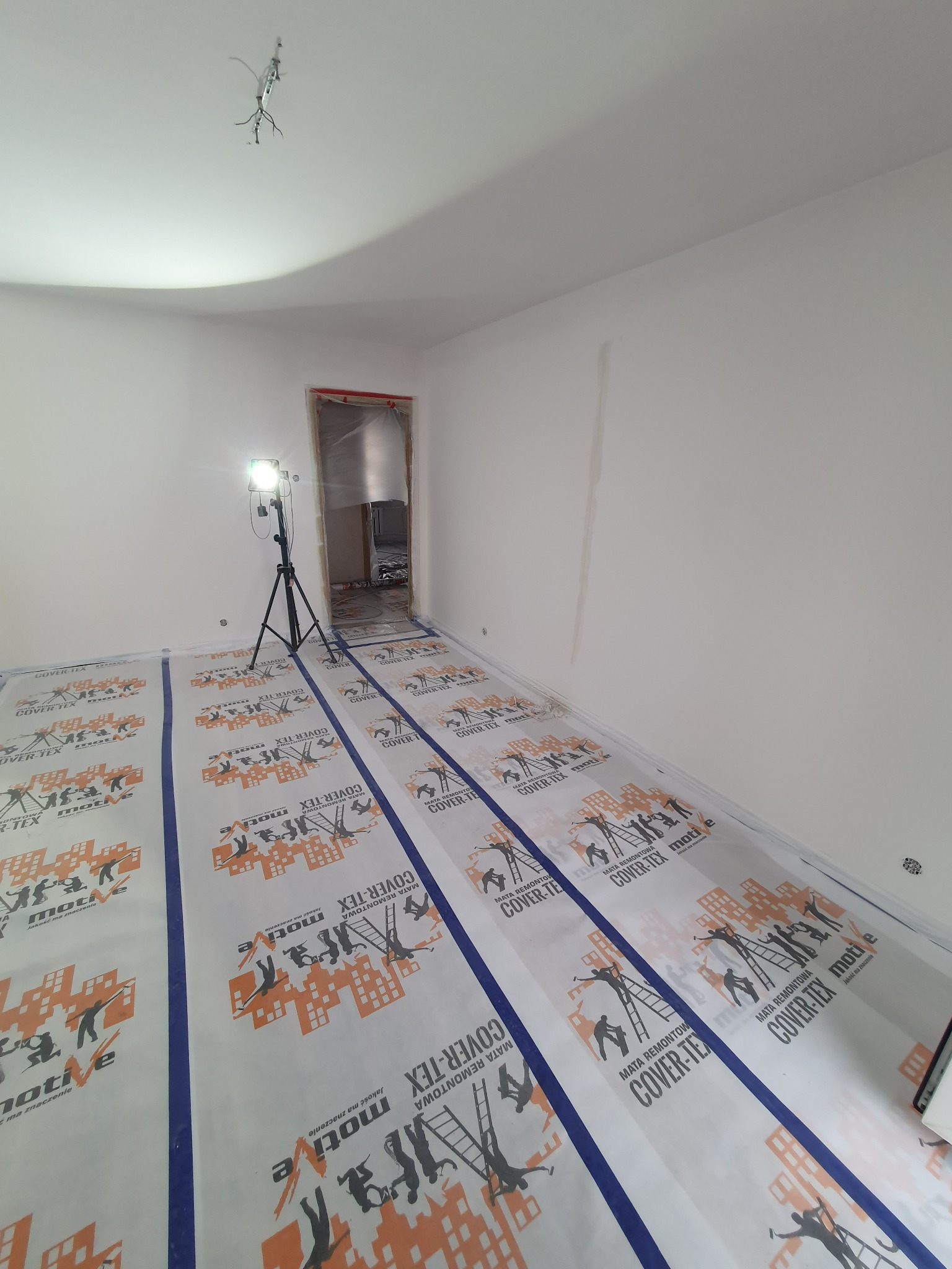 Pokój przygotowany do prac wykończeniowych, podłoga zabezpieczona folią ochronną z nadrukiem, widoczna lampa na statywie oświetlająca wnętrze, surowe ściany przed malowaniem.