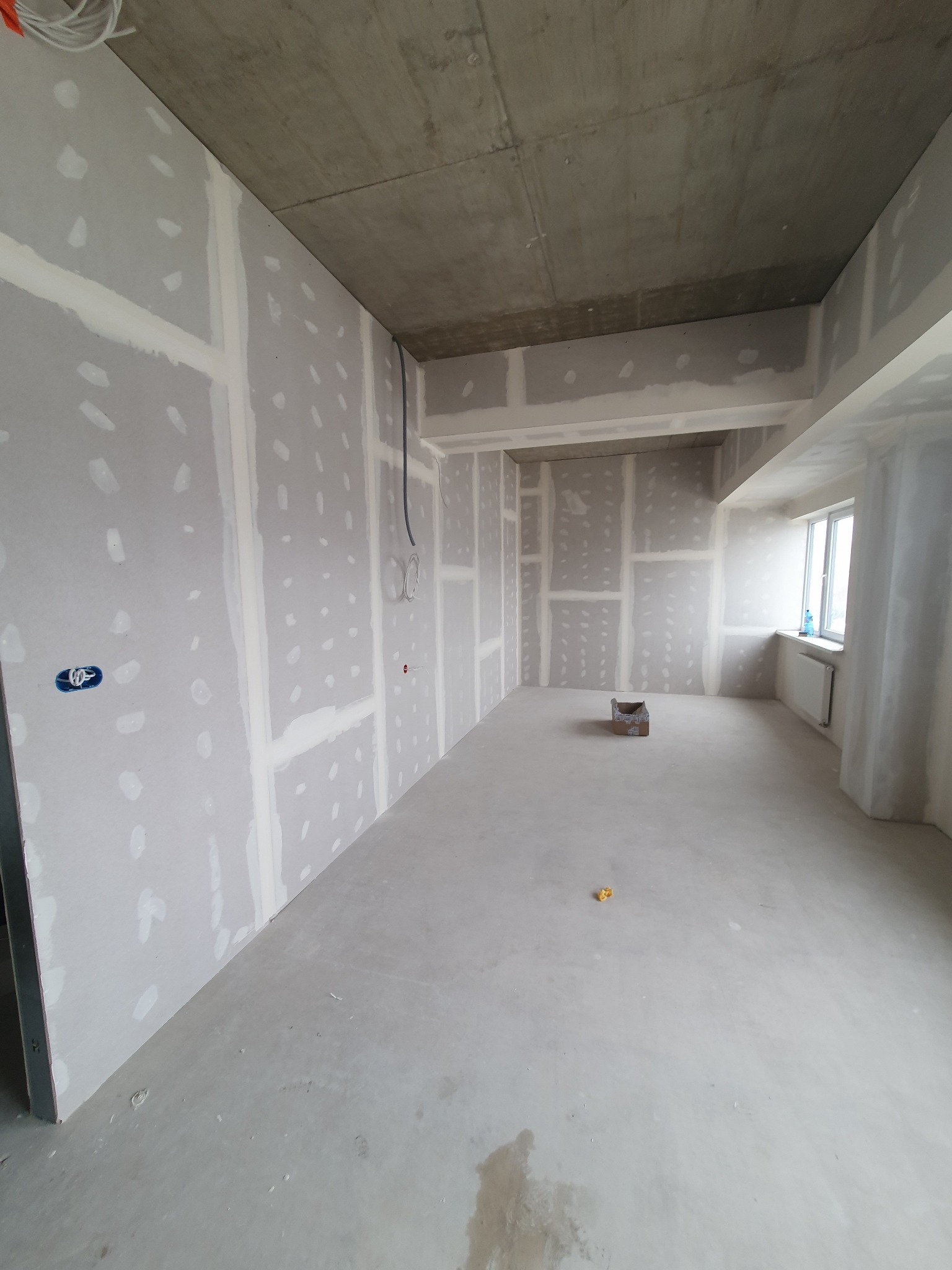 Pokój w surowym stanie po montażu płyt gipsowo-kartonowych, widoczne łączenia i otwory na gniazdka elektryczne, sufit betonowy.
