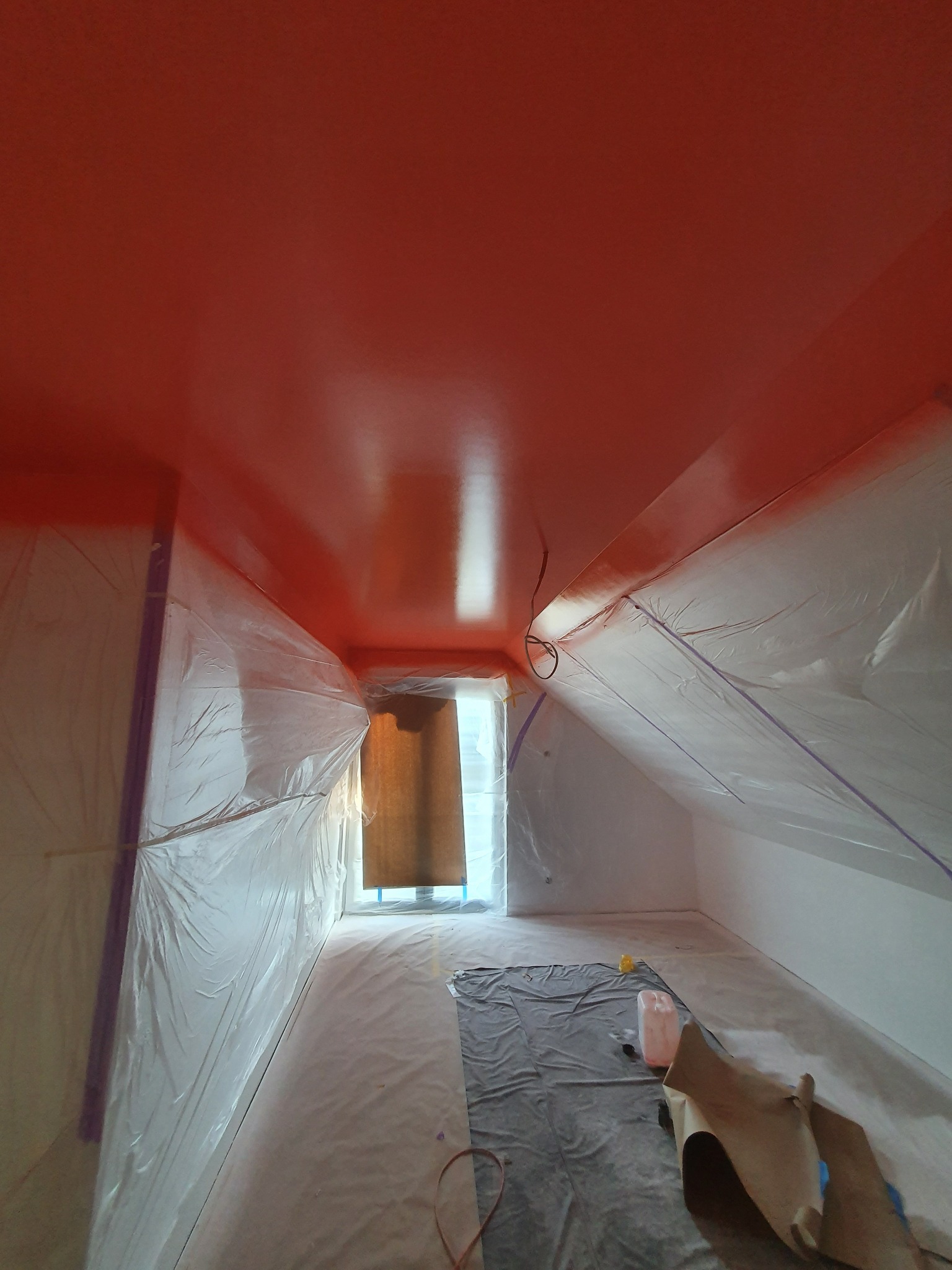Pokój na poddaszu zabezpieczony folią i taśmą malarską przed pomalowaniem sufitu na czerwono, widoczne narzędzia malarskie i wiadro z farbą.