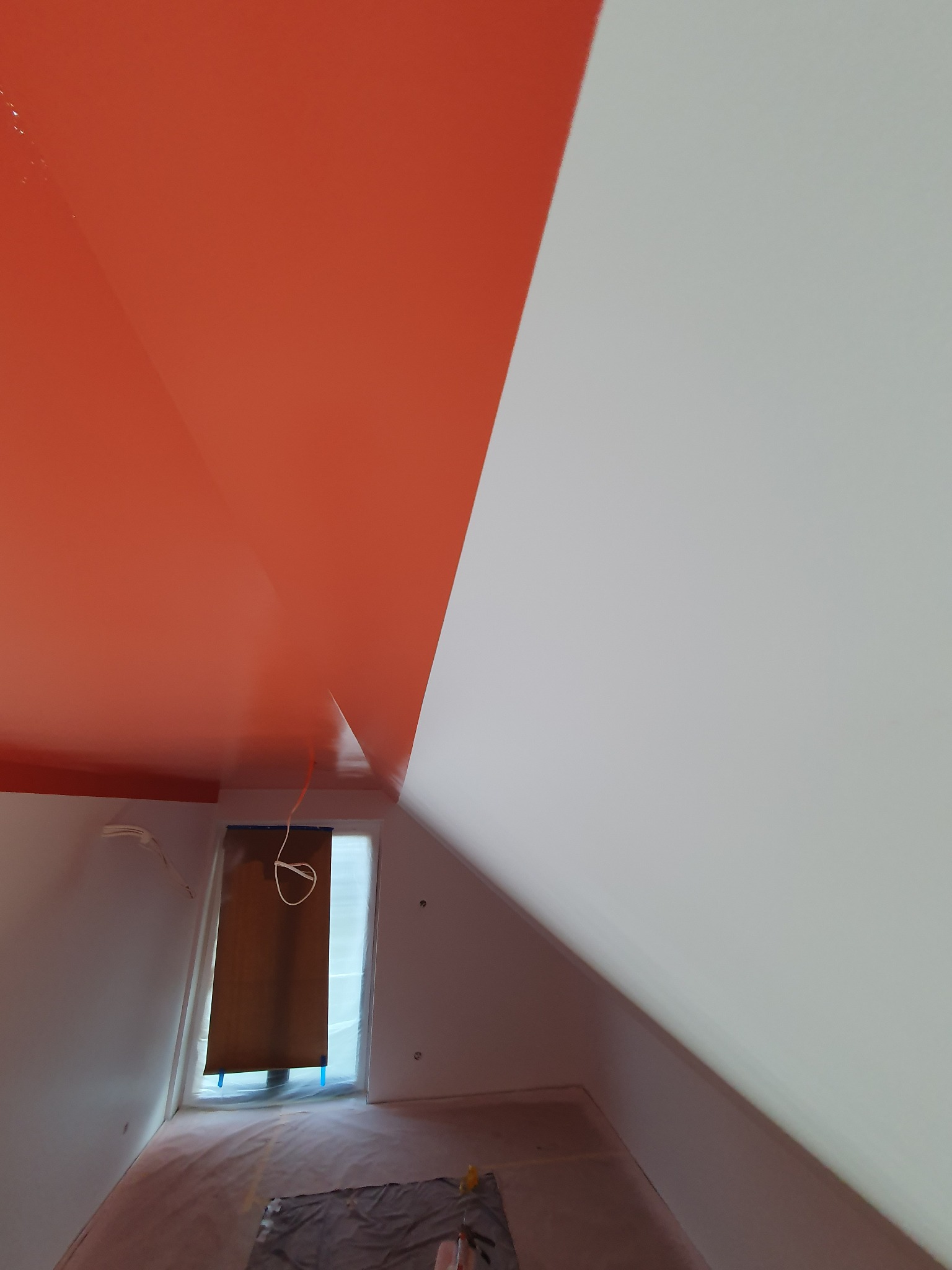 Pokój na poddaszu po częściowym pomalowaniu sufitu na pomarańczowo, druga część sufitu pomalowana na biało, okno zabezpieczone folią malarską, podłoga przykryta folią ochronną.