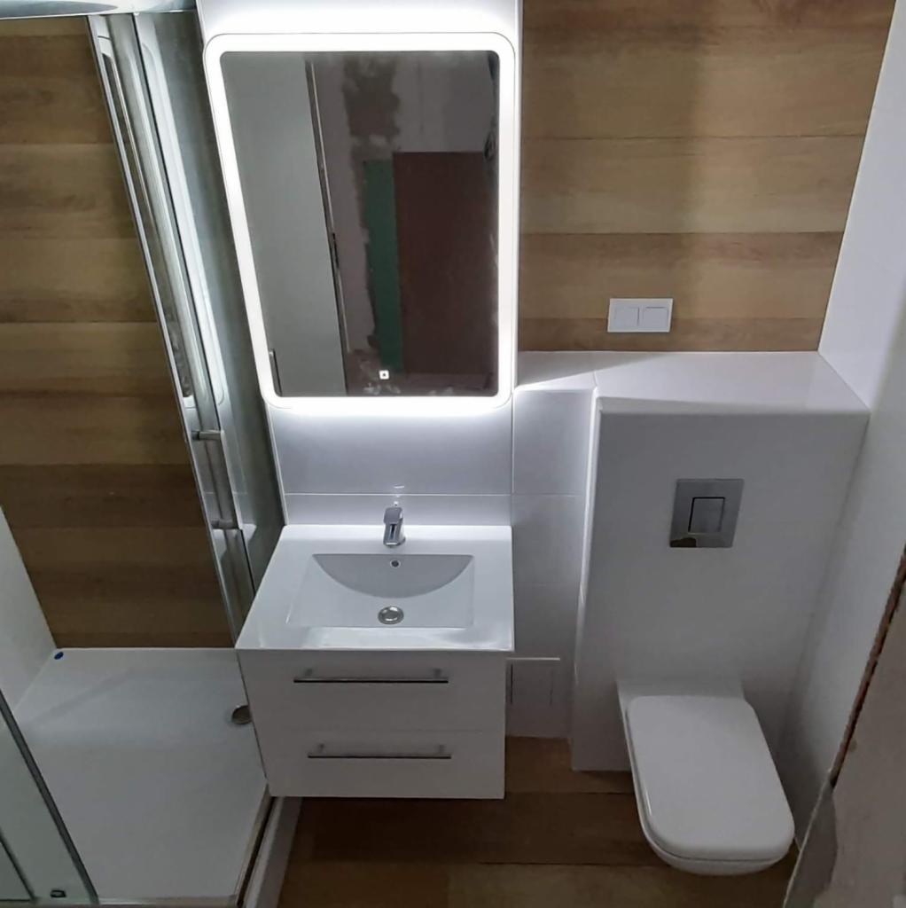 Nowoczesna łazienka po renowacji: biała armatura, drewniane panele ścienne i podświetlane lustro. Umywalka z szafką, toaleta podwieszana i kabina prysznicowa.