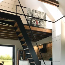 Loftowe wnętrze z metalowymi schodami z drewnianymi stopniami, antresolą z obrazem i widoczną kuchnią z kamienną ścianą.