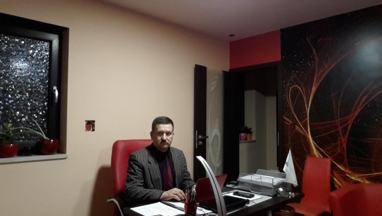 Mężczyzna w tweedowej marynarce siedzi przy biurku z laptopem, w tle okno z ozdobną szybą i abstrakcyjna tapeta.