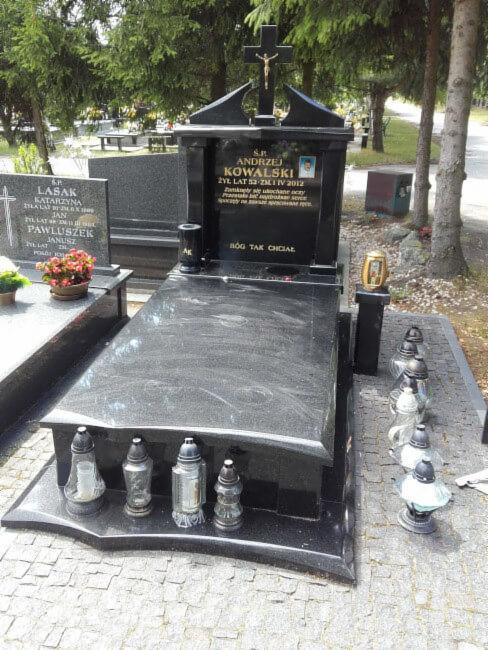 Czarny, granitowy pomnik z inskrypcjami i zdjęciem osoby zmarłej, ozdobiony zniczami i krzyżem, na tle zieleni cmentarza.