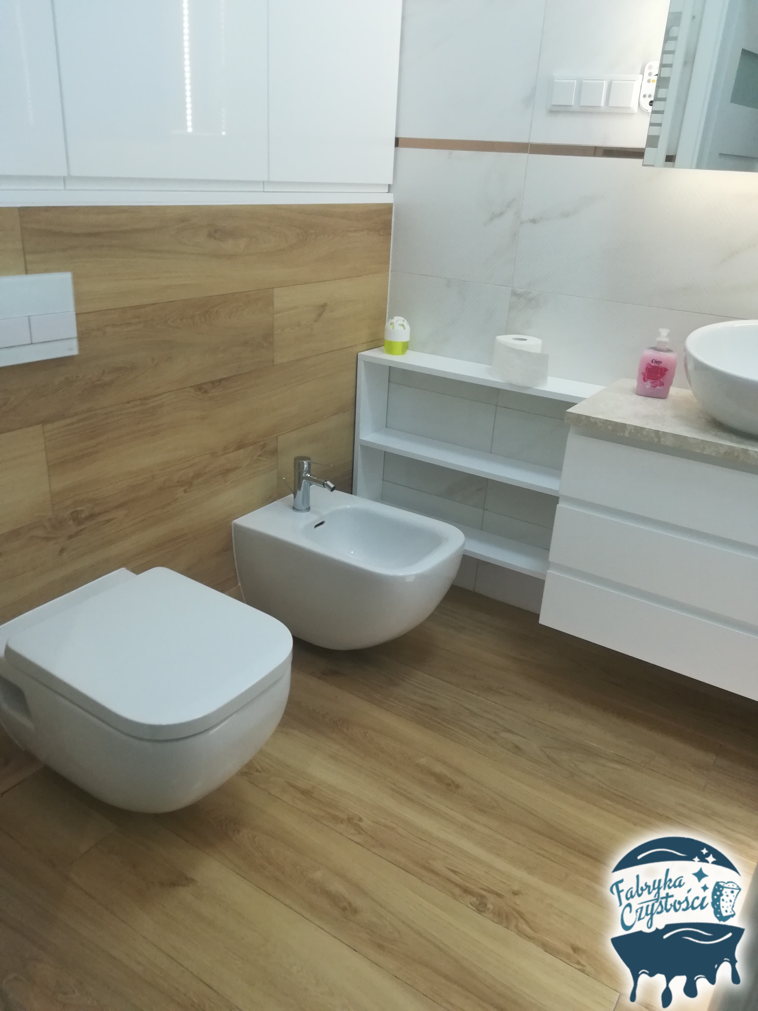 Nowoczesna łazienka z białymi sanitariatami (toaleta i bidet) oraz drewnopodobną podłogą, uzupełniona o białe szafki i półki. W rogu widoczne logo firmy sprzątającej.