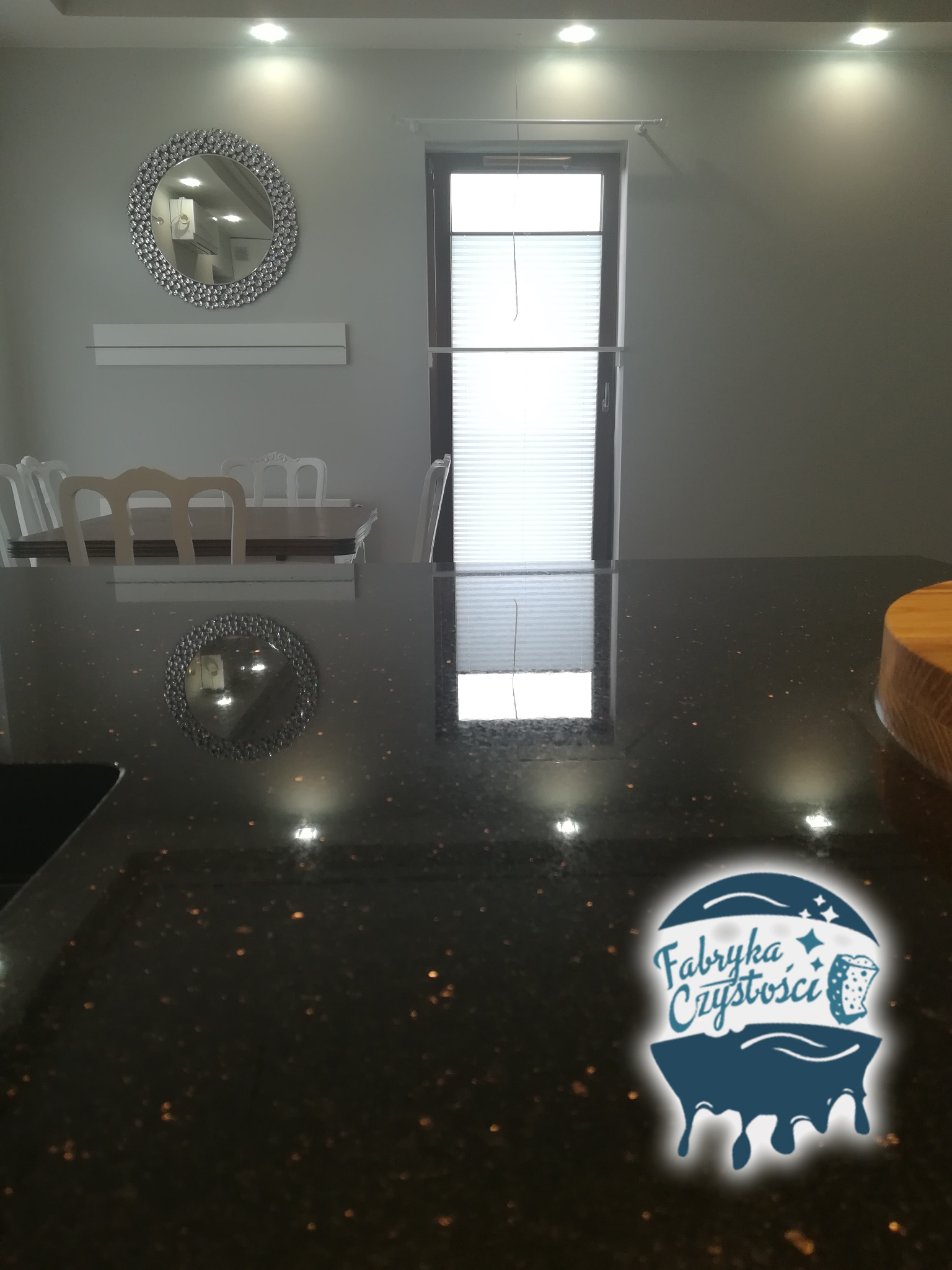 Błyszczący, czarny blat w kuchni z widocznym logiem firmy sprzątającej 'Fabryka Czystości', odbijającym się w lustrze na ścianie.