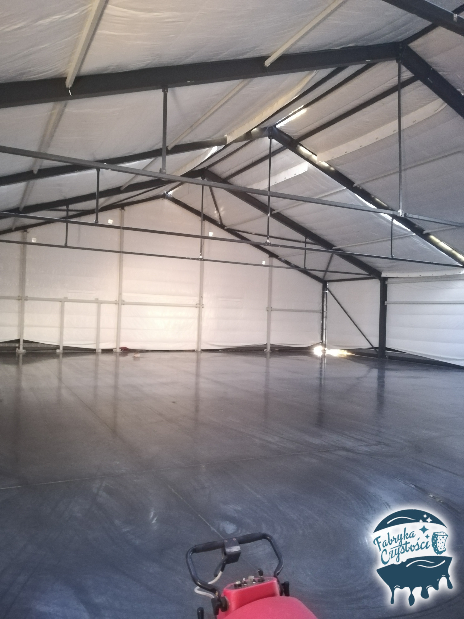 Wnętrze hali produkcyjnej z metalową konstrukcją dachu i białymi ścianami, czyszczone maszyną do mycia podłóg. Logo firmy sprzątającej w prawym dolnym rogu.