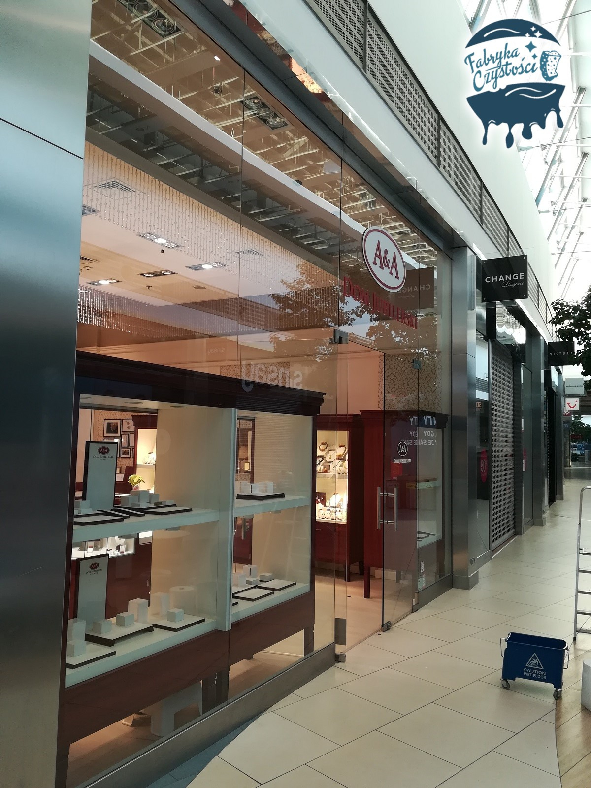 Wnętrze galerii handlowej z widoczną witryną sklepu jubilerskiego A&A po umyciu, z boku stoi niebieski wózek z napisem 'Caution Wet Floor', logo firmy sprzątającej Fabryka Czystości w górnym rogu.