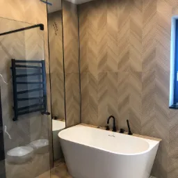 Nowoczesna łazienka z wolnostojącą białą wanną, czarną armaturą i ścianami wyłożonymi płytkami w jodełkę imitującymi drewno, widoczny szklany panel kabiny prysznicowej z czarnym grzejnikiem.