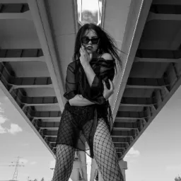 Czarno-białe zdjęcie kobiety w okularach przeciwsłonecznych, pozującej pod betonowym wiaduktem, z wiatrem we włosach.