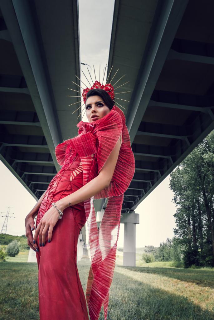 Kobieta w czerwonej, plisowanej sukni i koronie z promieniami pozuje pod betonowym wiaduktem, ujęcie z dołu.