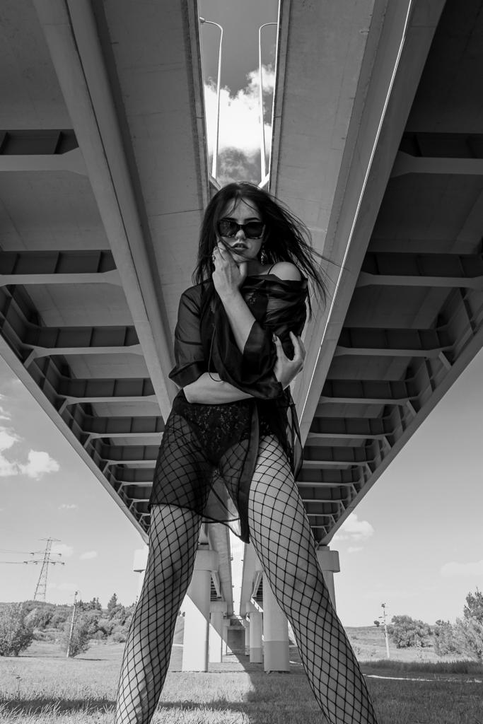 Czarno-białe zdjęcie kobiety w okularach przeciwsłonecznych, pozującej pod betonowym wiaduktem, z wiatrem we włosach.